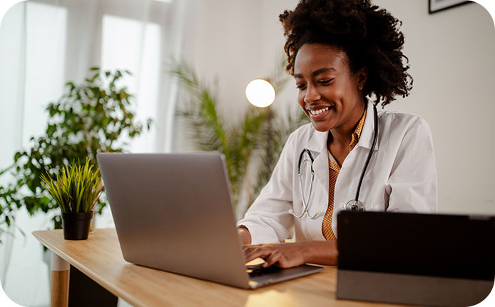 Attractive young African American woman doctor nurse practitioner assistant in hospital clinic office, typing on laptop.